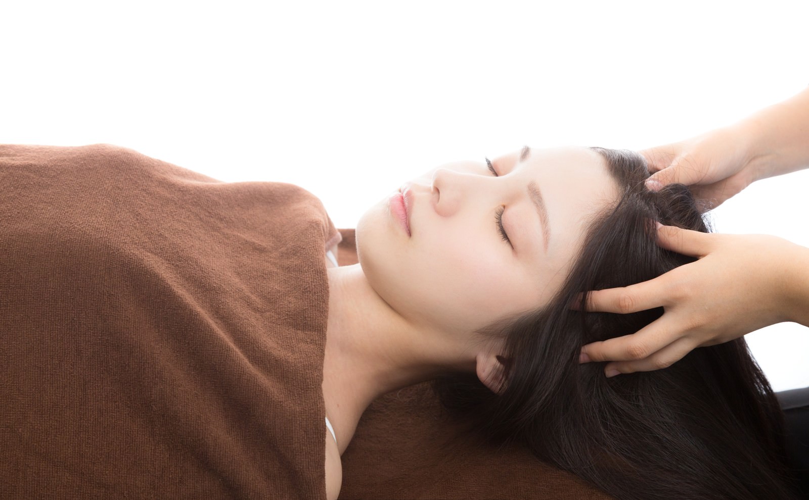 施術ベッドで頭部への整体施術を受ける30代女性。茶色のブランケットと明るい白背景で、首・頭まわりに女性施術者の手が添えられている